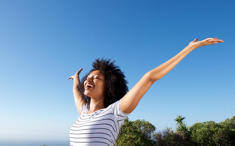 Portrait of young african woman standing outdoors with arms raised and laughing